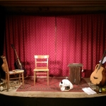 Bühne der Liedermacherlounge, roter Vorhang im Hintergrund; auf der Bühne stehen zwei Gitarren im Gitarrenstände,r zwei Sessel und ein Cajon