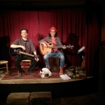 Reinauer im Duo mit Dixi auf der Bühne der Liedermacherlounge, Reinauer links sitzend mit Gitarre, Dixi rechts sitzend mit akustischem Bass