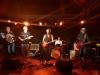 Bild der REINAUERAG live soielend,Bild ist in rotbraunes Licht gehalten, von linls nach rechts Stefan Wackerle an der Ziehharmonika, Hari Hittinger an der Gitarre und Mikro, REINAUER an der Gitarre und Mikro, Jens Gerö am Schlagzeug, Dixi am Bass